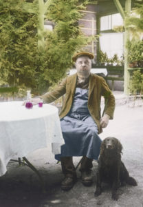 South Tyrol. Italy: Zecher with blue apron and dog in front of a South Tyrolean inn. Around 1925. Handcolored lantern slide.